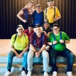 Theatercollectief Hoeksche Waard poseert in kleurrijke outfits en petten op een podium als voorbereiding op de voorstelling “De Jossen – Val en revival der saamhorigheid”.