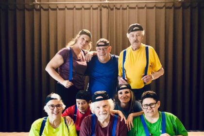 Theatercollectief Hoeksche Waard poseert in kleurrijke outfits en petten op een podium als voorbereiding op de voorstelling “De Jossen – Val en revival der saamhorigheid”.