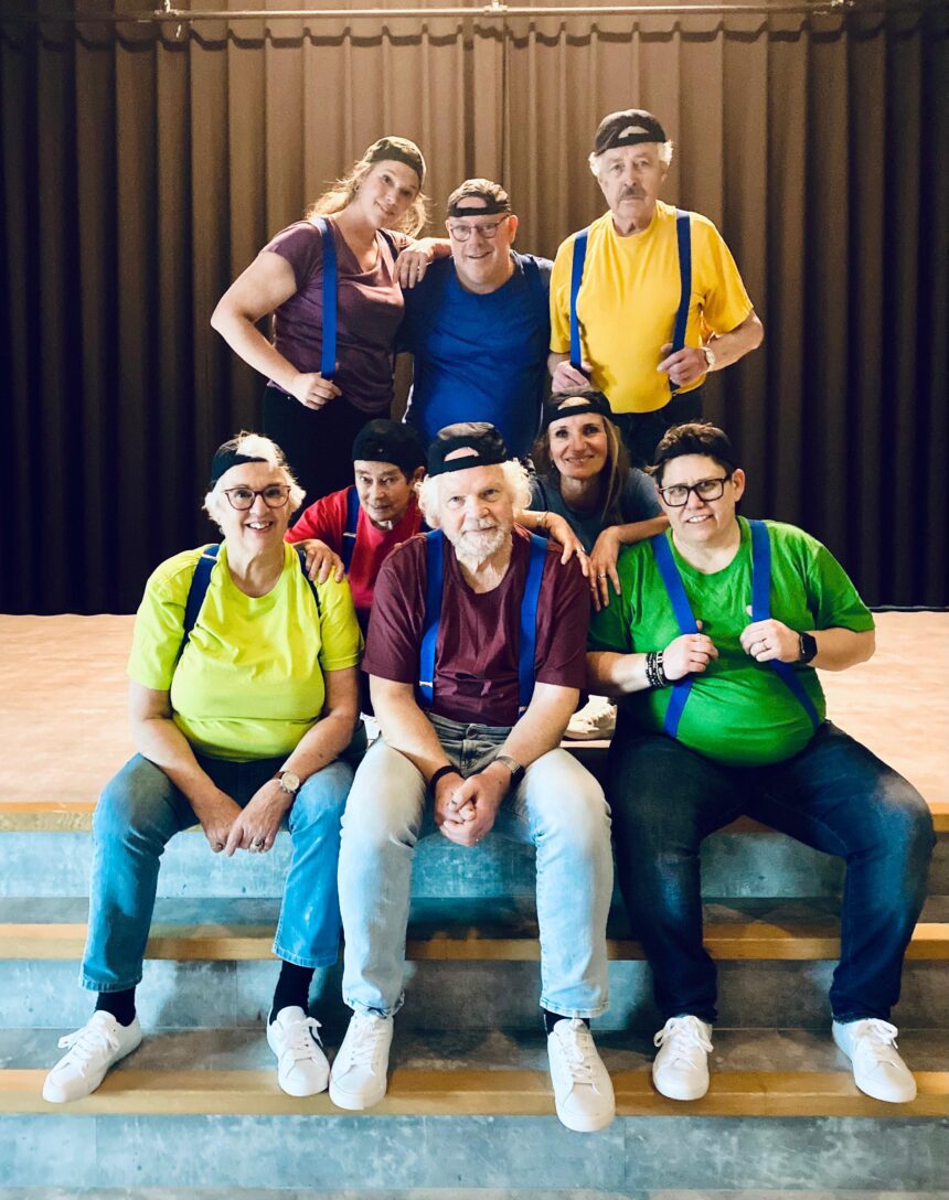 Theatercollectief Hoeksche Waard poseert in kleurrijke outfits en petten op een podium als voorbereiding op de voorstelling “De Jossen – Val en revival der saamhorigheid”.