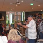 Leden van het Shantykoor De Hoeksche Waard treden op in de ontmoetingsruimte van Alerimus De Open Waard in Oud-Beijerland, begeleid door muzikanten, terwijl bewoners aandachtig luisteren en meezingen.