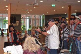 Leden van het Shantykoor De Hoeksche Waard treden op in de ontmoetingsruimte van Alerimus De Open Waard in Oud-Beijerland, begeleid door muzikanten, terwijl bewoners aandachtig luisteren en meezingen.